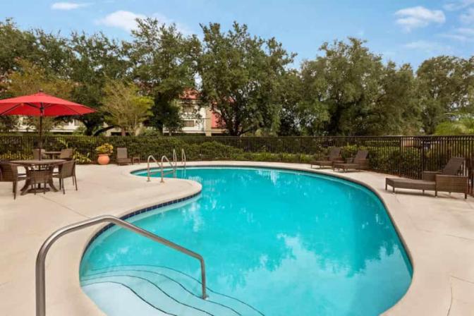 Elegant outdoor pool at Ramada Suites, complete with pristine water, ample lounge chairs, palm trees, and shaded areas under umbrellas, set against the backdrop of the hotel’s contemporary architecture.
