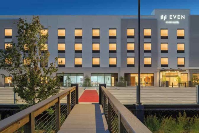 EVEN Hotel's exterior at dusk featuring a welcoming red-carpeted boardwalk entrance, warm interior lighting through tall windows, and minimalist landscaping around a contemporary façade.