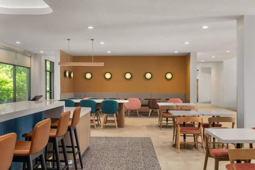 Colorful dining area at EVEN Hotel showcasing a variety of modern chairs, high-top bar seating, and round wall lights against a mustard accent wall, designed for casual social dining.