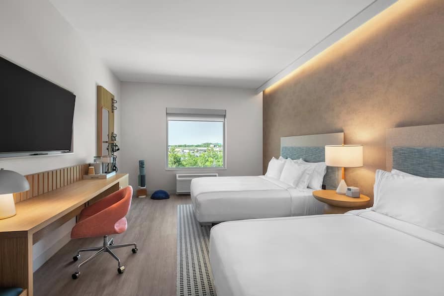 Spacious EVEN Hotel guest room showcasing two queen beds with upholstered headboards, a sleek wood-accented desk with a coral swivel chair, a flat-screen TV, and a large window with views of greenery.