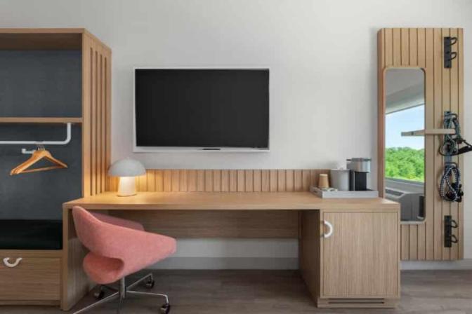 Functional in-room workspace at EVEN Hotel featuring a wood desk with a modern coral chair, flat-screen TV, coffee station, and full-length mirror with resistance bands mounted beside a wardrobe nook.