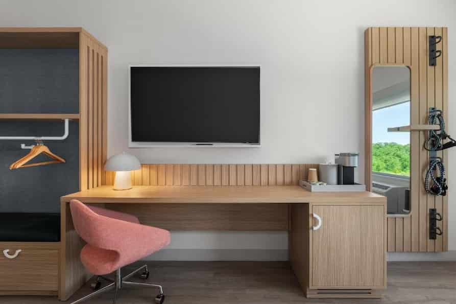Functional in-room workspace at EVEN Hotel featuring a wood desk with a modern coral chair, flat-screen TV, coffee station, and full-length mirror with resistance bands mounted beside a wardrobe nook.