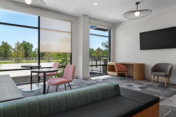 Cozy lounge at EVEN Hotel featuring a sleek black-back sofa, coral accent chairs, a round table, and a flat-screen TV mounted above a compact wooden desk, all framed by tall windows.