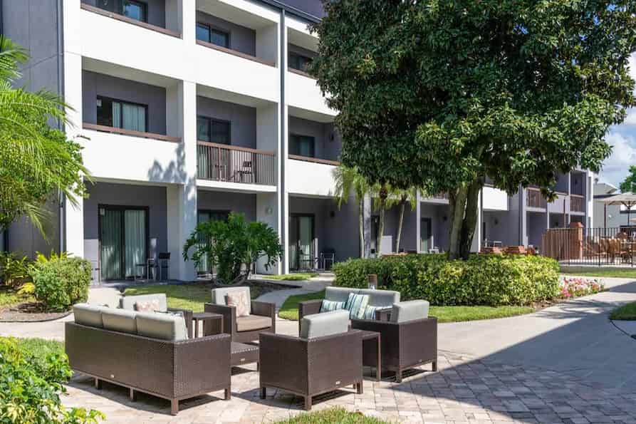 Outdoor patio area at the Courtyard by Marriott hotel featuring comfortable wicker seating with cushions, surrounded by lush greenery and the exterior of the hotel building in the background.
