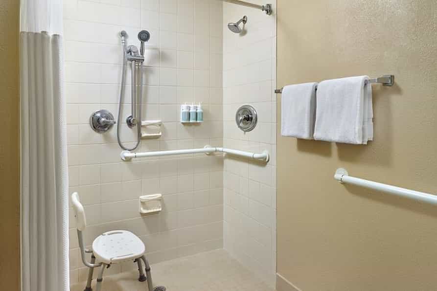 Spacious accessible bathroom in the Courtyard by Marriott hotel, featuring a walk in shower with grab bars, a shower chair, and wall mounted toiletries and towels neatly placed on a towel rack.