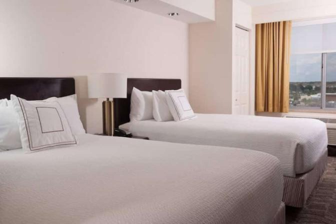 Hotel room at the SpringHill Suites by Marriott featuring two queen beds with white bedding, a modern side table lamp and a large window overlooking a town center.