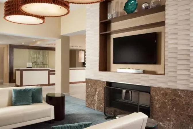 Modern hotel lobby featuring a cozy seating area, flat-screen TV, and fireplace with reception desk in the background.
