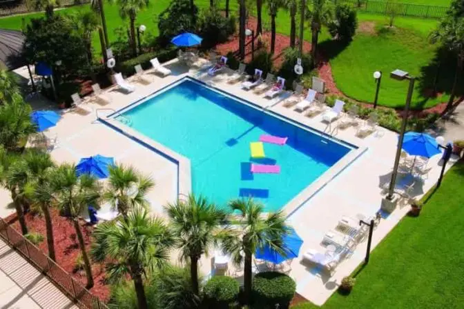 Aerial view of an outdoor swimming pool surrounded by palm trees, lounge chairs with blue umbrellas, and colorful pool floats.