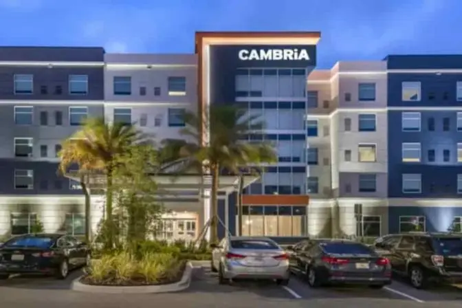 Exterior view of Cambria Orlando International Airport Hotel, featuring a modern entrance with warm lighting and tall palm trees displayed against an evening sky.