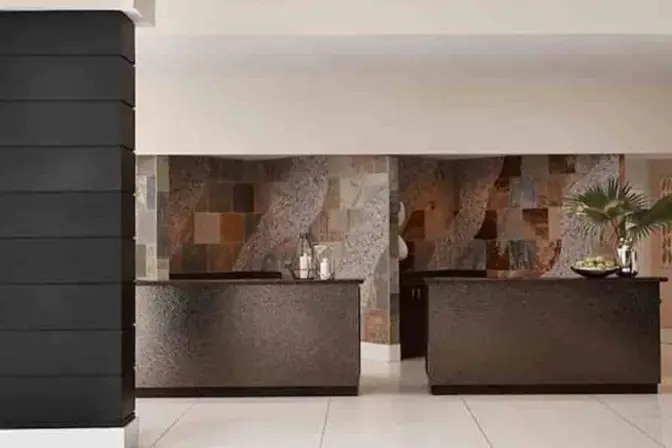 Hotel front desk at Renaissance Orlando Airport with textured black check-in counters, a backdrop of natural stone tiles in earth tones, and minimalist decor including candles and tropical greenery.