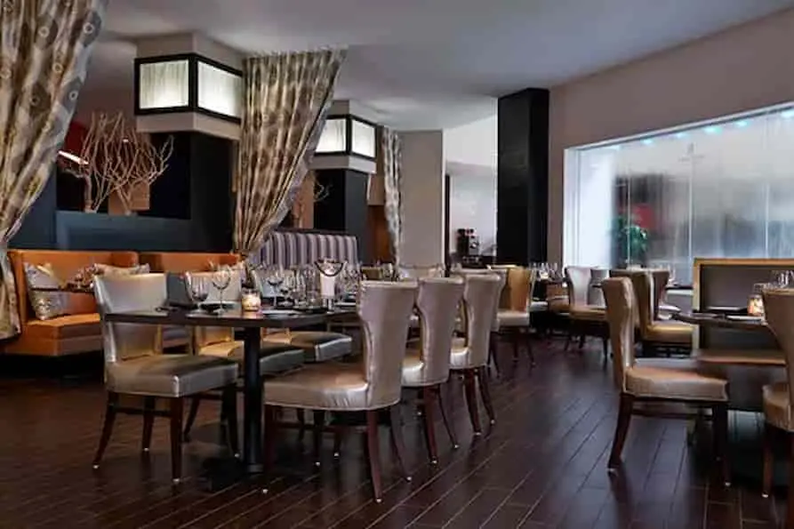 Upscale restaurant at Renaissance Orlando Airport Hotel featuring dark wood flooring, contemporary dining tables, metallic leather chairs, cozy booth seating, and ambient lighting with flowing curtains.