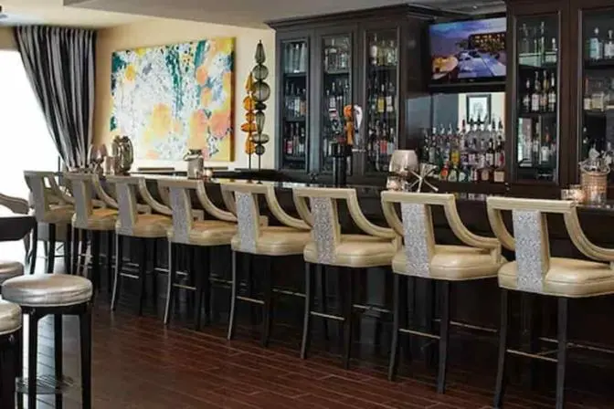 Renaissance Orlando Airport Hotel bar featuring high-top stools with silver upholstery, a dark wood bar, backlit glass shelves stocked with spirits, and modern artwork on a yellow accent wall.