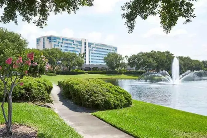 Scenic outdoor view of Renaissance Orlando Airport Hotel with a winding walking path beside a landscaped pond, lush greenery, blooming trees, and a water fountain in front of the modern hotel façade.