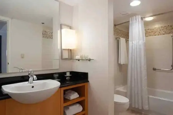 Bathroom at Renaissance Orlando Airport Hotel with a vessel sink, granite countertop, open shelving, lighted vanity mirror, and a tub-shower combo with safety rails and mosaic tile accents.