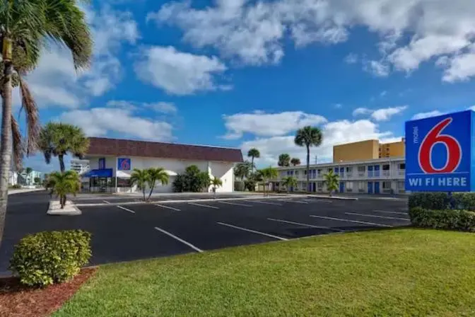 Exterior view of Motel 6 Cocoa Beach with palm trees and a spacious parking lot on a sunny day.