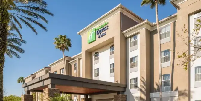 Exterior view of Holiday Inn Express Orlando International Airport Hotel, featuring a bright terracotta and beige building with tall palm trees displayed against a bright afternoon sky.