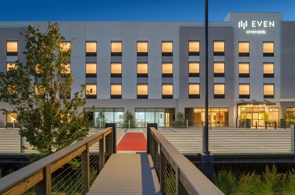EVEN Hotel's exterior at dusk featuring a welcoming red-carpeted boardwalk entrance, warm interior lighting through tall windows, and minimalist landscaping around a contemporary façade.