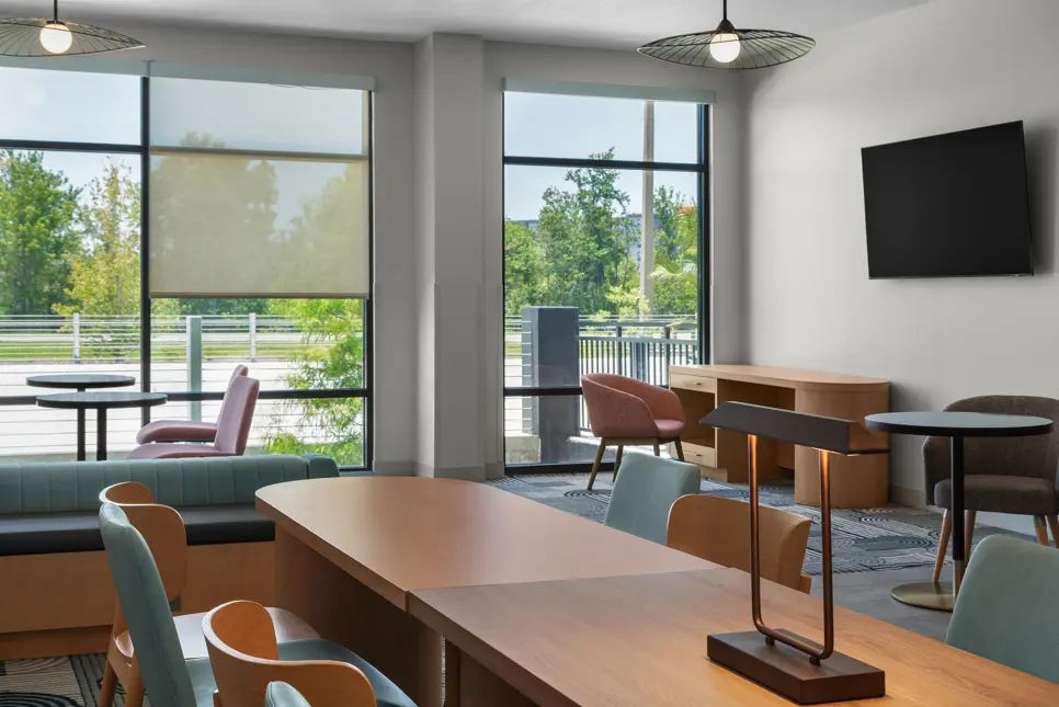Cozy lounge at EVEN Hotel featuring a sleek black-back sofa, coral accent chairs, a round table, and a flat-screen TV mounted above a compact wooden desk, all framed by tall windows.