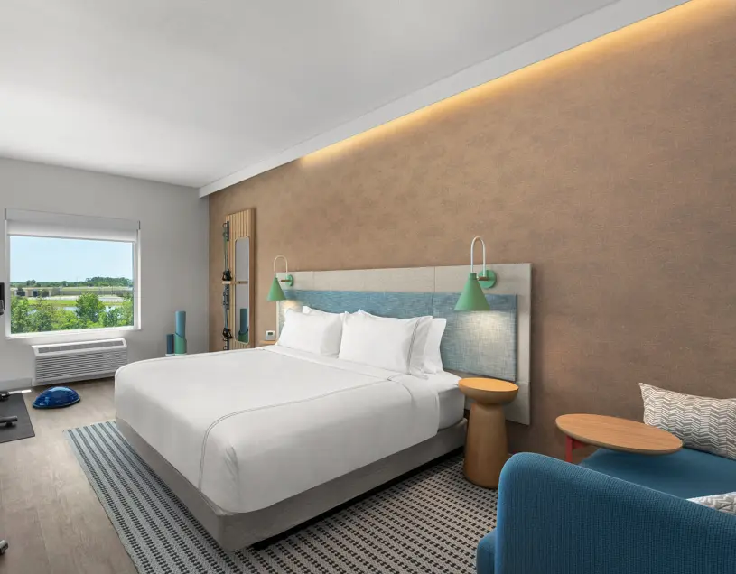 EVEN Hotel's king guest room featuring a modern upholstered headboard with green wall-mounted lamps, crisp white bedding, a blue lounge chair with side tables, in-room fitness equipment, and a large window with scenic views.