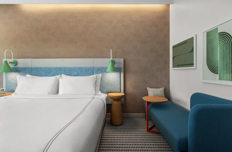 EVEN Hotel's king guest room with a textured accent wall, green wall-mounted lamps, crisp white linens, a blue upholstered bench with side table, and modern green geometric artwork above the seating area.