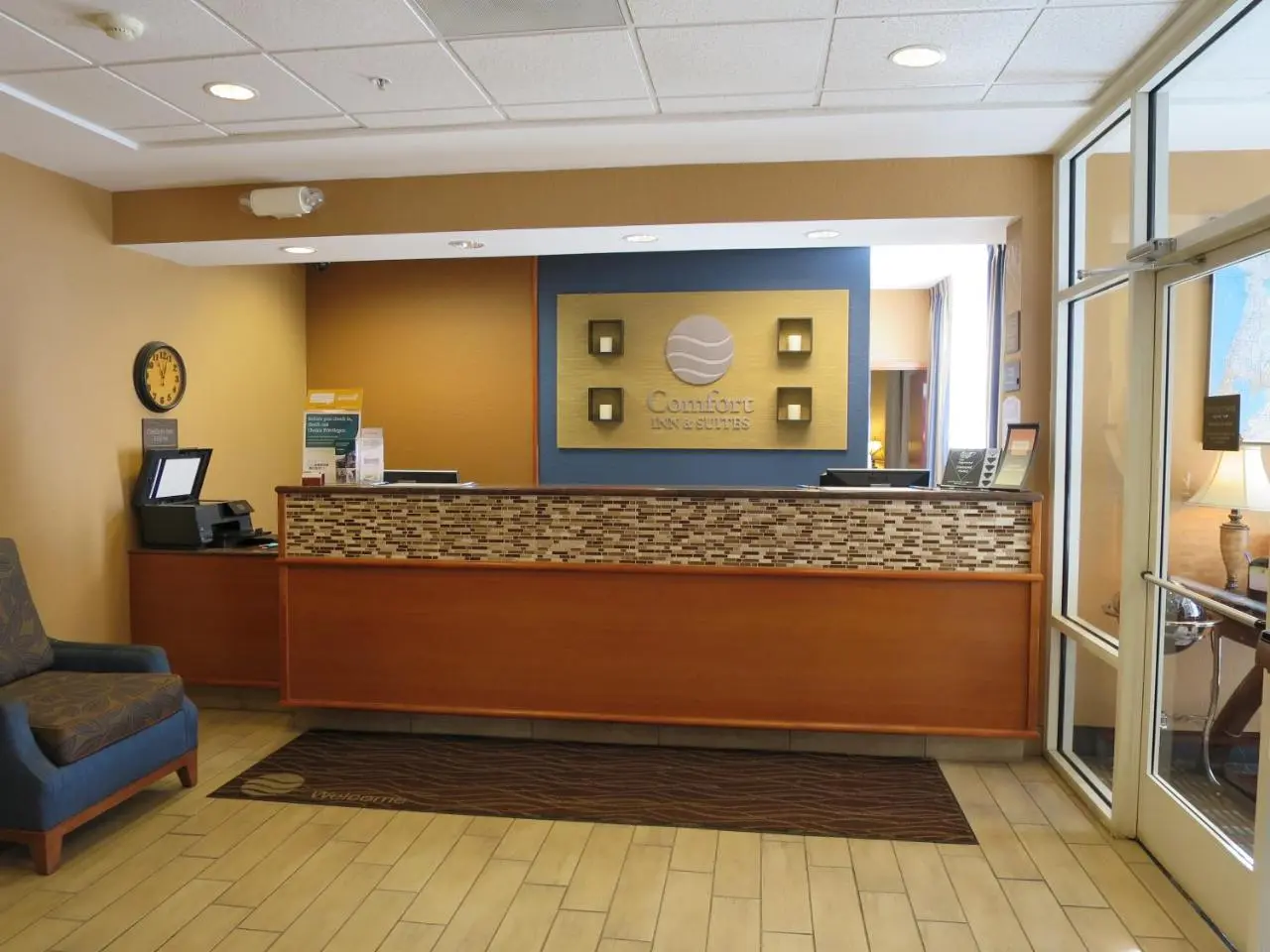 Reception area at Comfort Inn & Suites Sanford hotel showcasing a stylish front desk, warm lighting, and a comfortable blue armchair in the lobby.