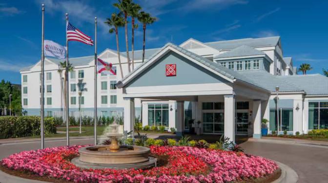Exterior view of Hilton Garden Inn Orlando Airport Hotel during the day, featuring the main entrance with a water fountain surrounded by pink flowers and palm trees
