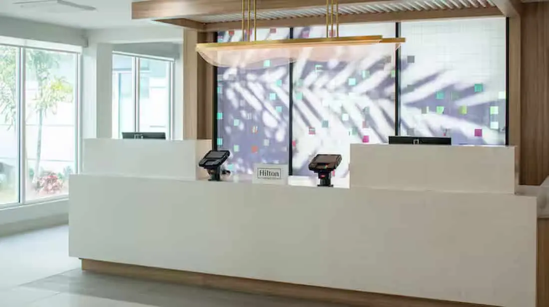 Lobby reception area of Hilton Garden Inn Orlando Airport Hotel featuring a modern white front desk, sleek lighting fixture, and decorative frosted glass backdrop with colorful accents, surrounded by bright natural light from large windows.