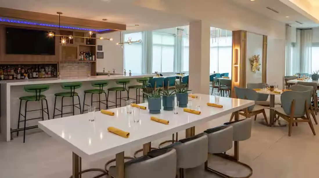 Modern dining area at Hilton Garden Inn Orlando Airport featuring a sleek bar with green stools, contemporary lighting, and tables set with yellow napkins and potted plants in a bright, airy space.
