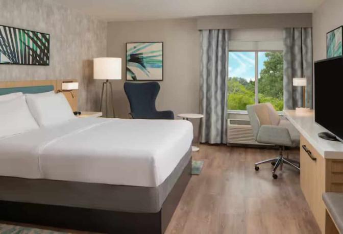 Modern hotel room with a king-sized bed, an armchair, a work desk, and a TV, with large windows providing natural light.