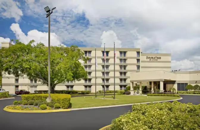 Exterior view of DoubleTree by Hilton Hotel Orlando East-UCF Area featuring a multi-story beige building with a covered entrance, landscaped greenery, and flagpoles under a bright blue sky with scattered clouds.