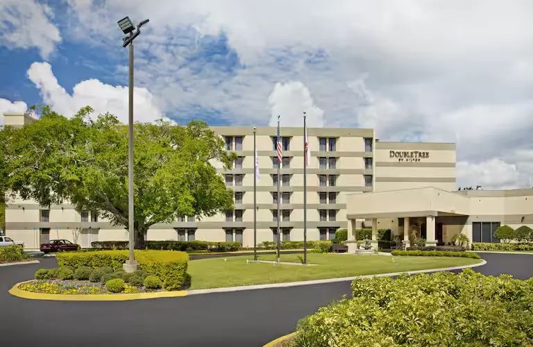 Exterior view of DoubleTree by Hilton Hotel Orlando East-UCF Area featuring a multi-story beige building with a covered entrance, landscaped greenery, and flagpoles under a bright blue sky with scattered clouds.