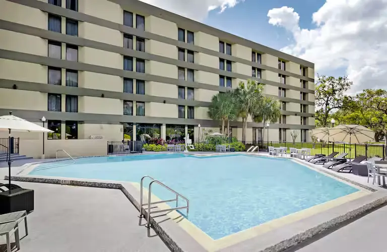 Outdoor pool area at DoubleTree by Hilton Hotel Orlando East-UCF Area featuring a spacious swimming pool surrounded by lounge chairs, umbrellas, and palm trees against the hotel’s exterior backdrop.