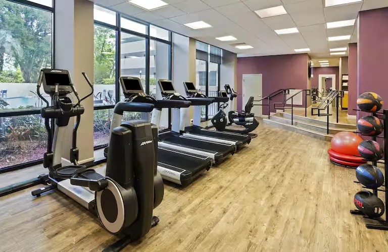Fitness center at DoubleTree by Hilton Hotel Orlando East-UCF Area featuring treadmills, ellipticals, stationary bikes, and strength training equipment with floor-to-ceiling windows overlooking the outdoor pool.