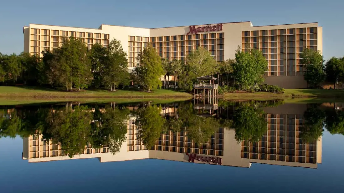 Exterior view of the Marriott Orlando Airport Lakeside Hotel featuring modern architecture surrounded by lush greenery, reflected in a calm lake under a clear blue sky.