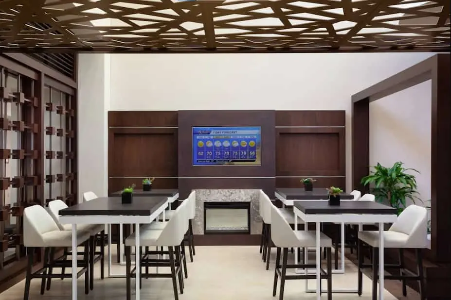 Hotel lounge area at Marriott Lakeside with high-top tables, white chairs, and a large screen displaying the weather forecast.