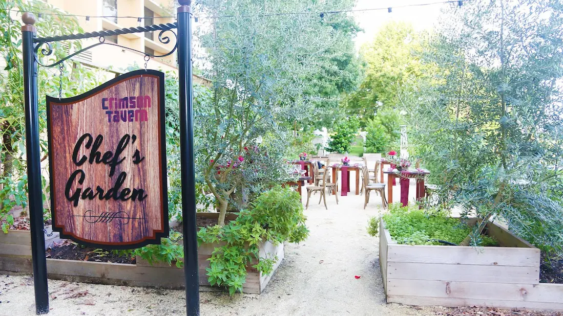 Outdoor dining experience at Crimson Tavern Chef's Garden at the Marriott Lakeside Orlando International Airport Hotel, showcasing a wooden sign and tables nestled among plants and trees.
