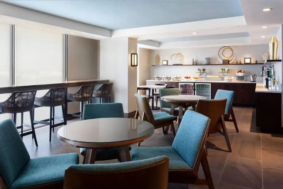 Stylish lounge featuring comfortable seating, bar stools at a counter, and a variety of food and drink options displayed in the background.