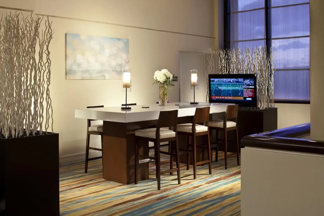 Stylish hotel lounge featuring a communal high-top table, bar stools, ambient lighting, and a TV for entertainment.