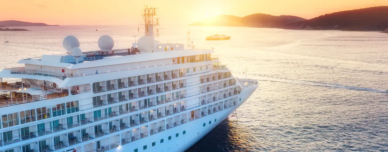 Large white cruise ship sailing at sunset near the coast, surrounded by calm blue sea and distant hills.