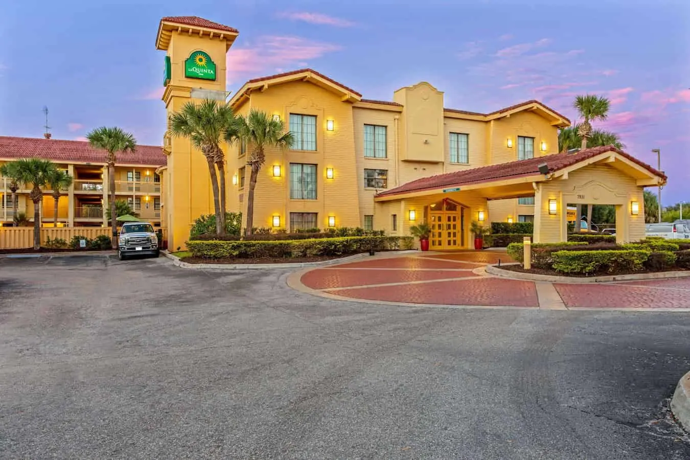 Exterior view of La Quinta Inn by Wyndham Orlando Airport West with palm trees and covered entrance at sunset.