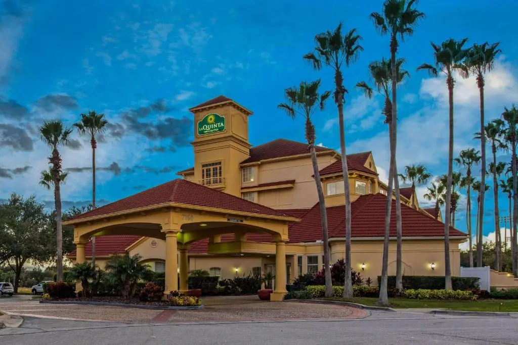 Exterior view of La Quinta Inn & Suites Orlando International Airport Hotel, featuring a grand entrance with tall palm trees displayed against an afternoon sky.
