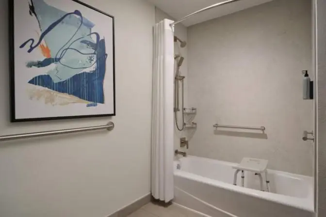 Accessible hotel bathroom with a bathtub, grab bars, handheld showerhead, and a shower chair inside the tub.