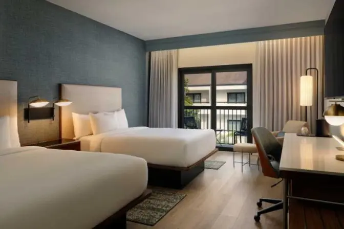 Hotel room at the Courtyard by Marriott featuring two queen beds with white bedding, a work desk with a lamp and chair, a flat-screen TV, and a tan armchair near the window.