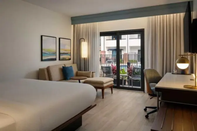 A modern hotel room with a king bed, a beige sofa and small table, a desk with a chair and lamp, and sliding glass doors leading to a balcony with outdoor seating.