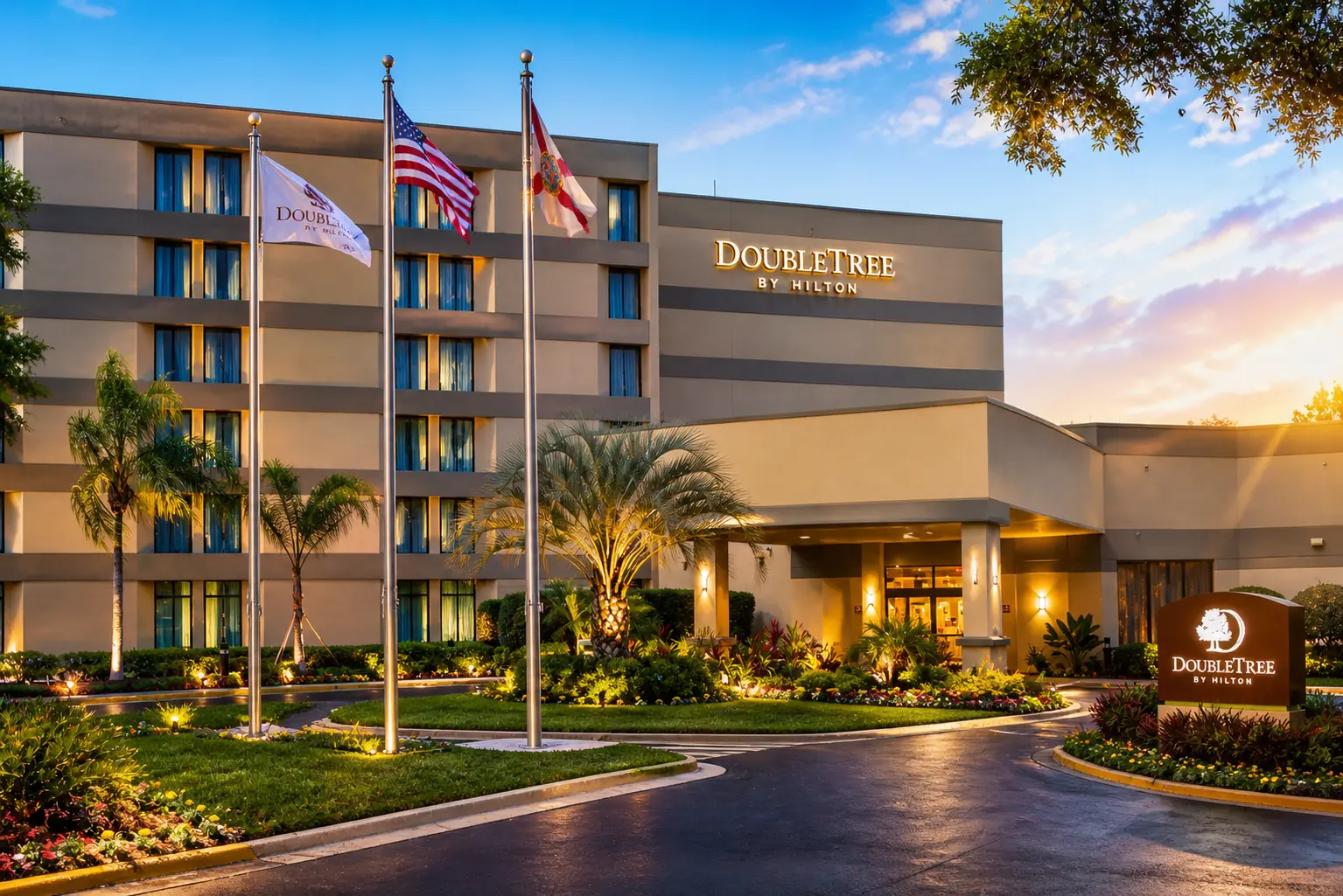 Exterior view of DoubleTree by Hilton hotel featuring a multi-story beige building with a covered entrance, landscaped greenery, palm trees, and flagpoles under a bright blue sky at sunset.
