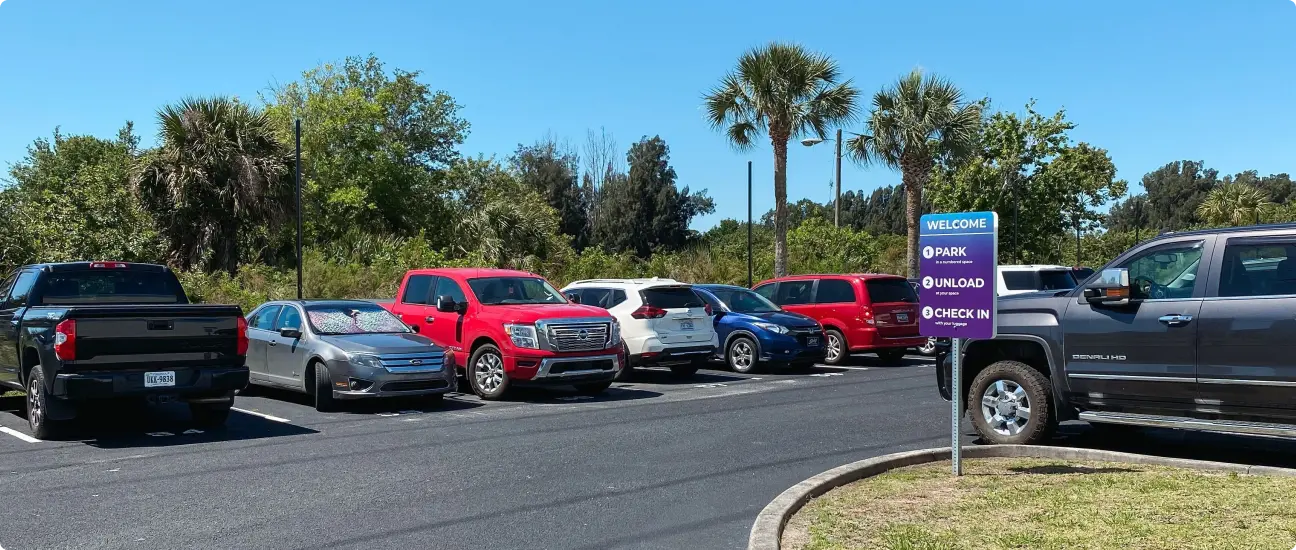 Parking area at Go Port Plaza with various parked cars, a welcome sign providing parking instructions, and palm trees in the background under a clear blue sky.